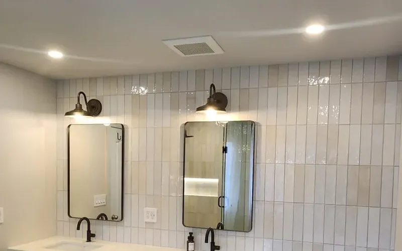 Bathroom with vertical white glazed tile, two black-framed mirrors under black gooseneck sconces, pot lights and a rectangular grille in the white ceiling above the mirrors