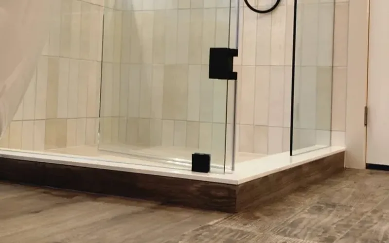 Low view of a glass shower on a white base, wood-look floor tile running up to the base, white vertical wall tile behind