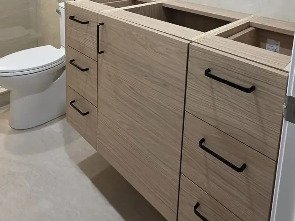 Large pale floor tile in a finished bathroom, a toilet on the left and a wall-hung oak vanity with black pulls, its top not yet installed