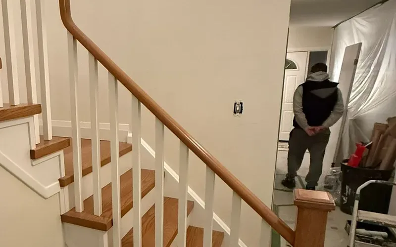 Refinished oak stair with white spindles; behind it a hallway sealed off with clear plastic sheeting, the floor covered with protection paper and a worker walking toward the front door