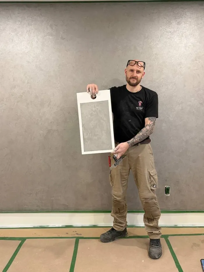 Art Style plasterer in a black company T-shirt holding a framed grey sample board and a trowel in front of a finished grey plaster wall, floor covered in paper