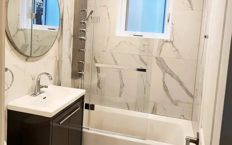 Small bathroom with a dark vanity, round mirror, marble-look tile and a bathtub with a glass screen below a window