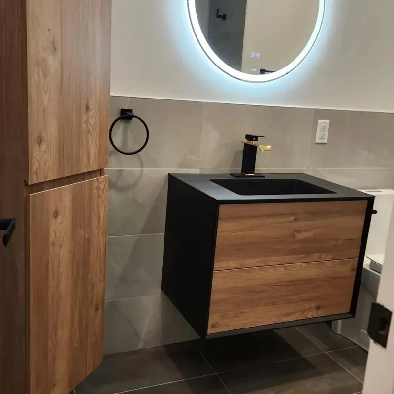 Wall-hung wood and black vanity under a round backlit mirror, with a tall wood cabinet beside it and grey tile walls