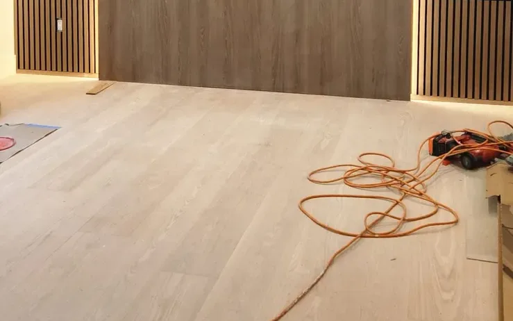 Light oak plank floor in front of a wall of wood panels and slatted panels with LED strips glowing at their edges, an orange extension cord and power tools still lying on the floor