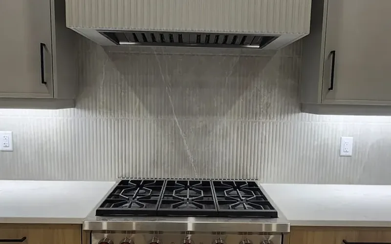 Ribbed porcelain backsplash rising between grey upper cabinets to a fluted range hood above a gas range