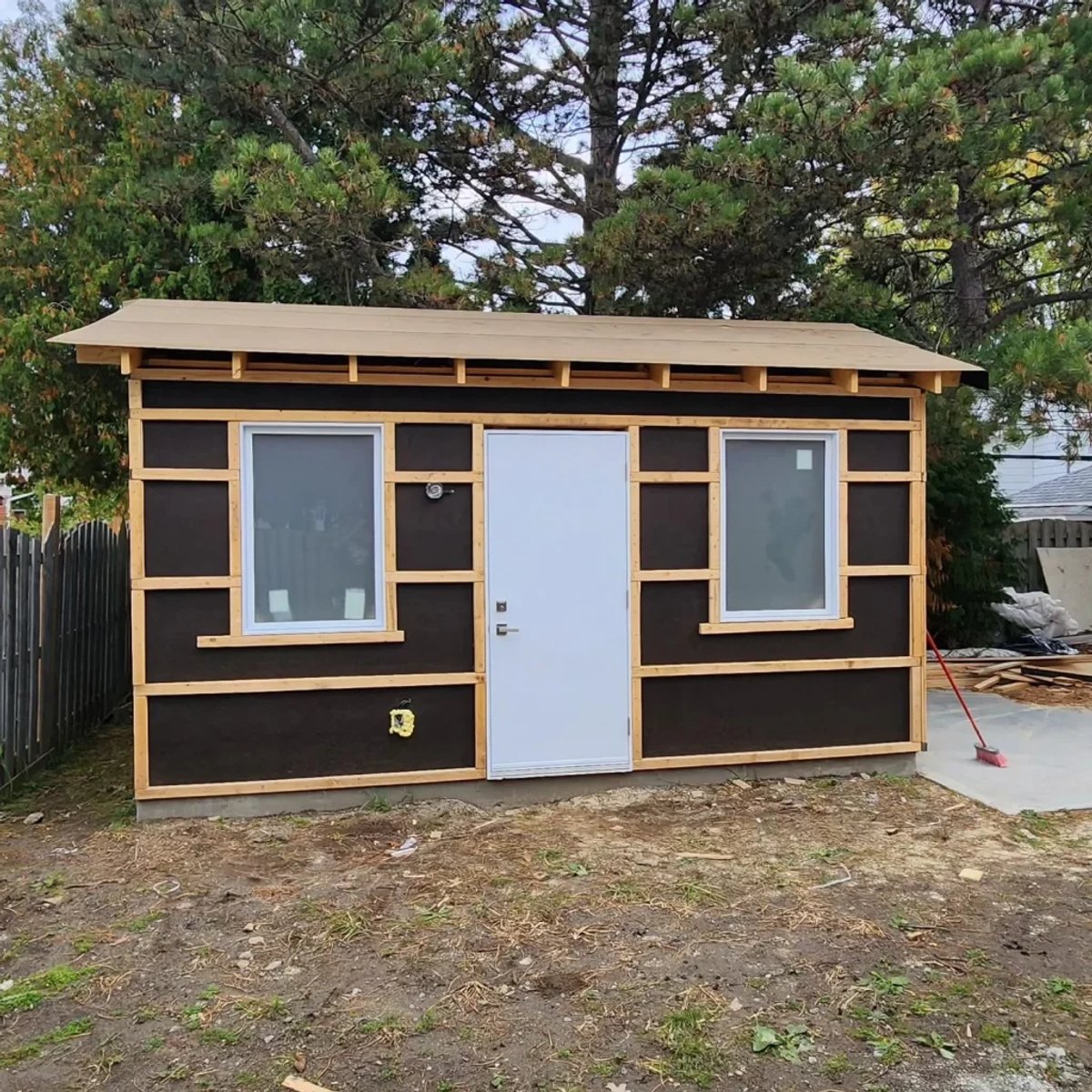 Small backyard building with dark sheathing, wood trim strips, two windows and a white door under pine trees