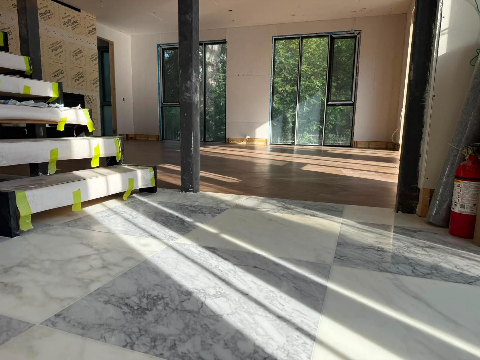 Grey and white marble checkerboard floor beside steel stair treads wrapped in white board and yellow tape, with a wood floor and tall windows beyond