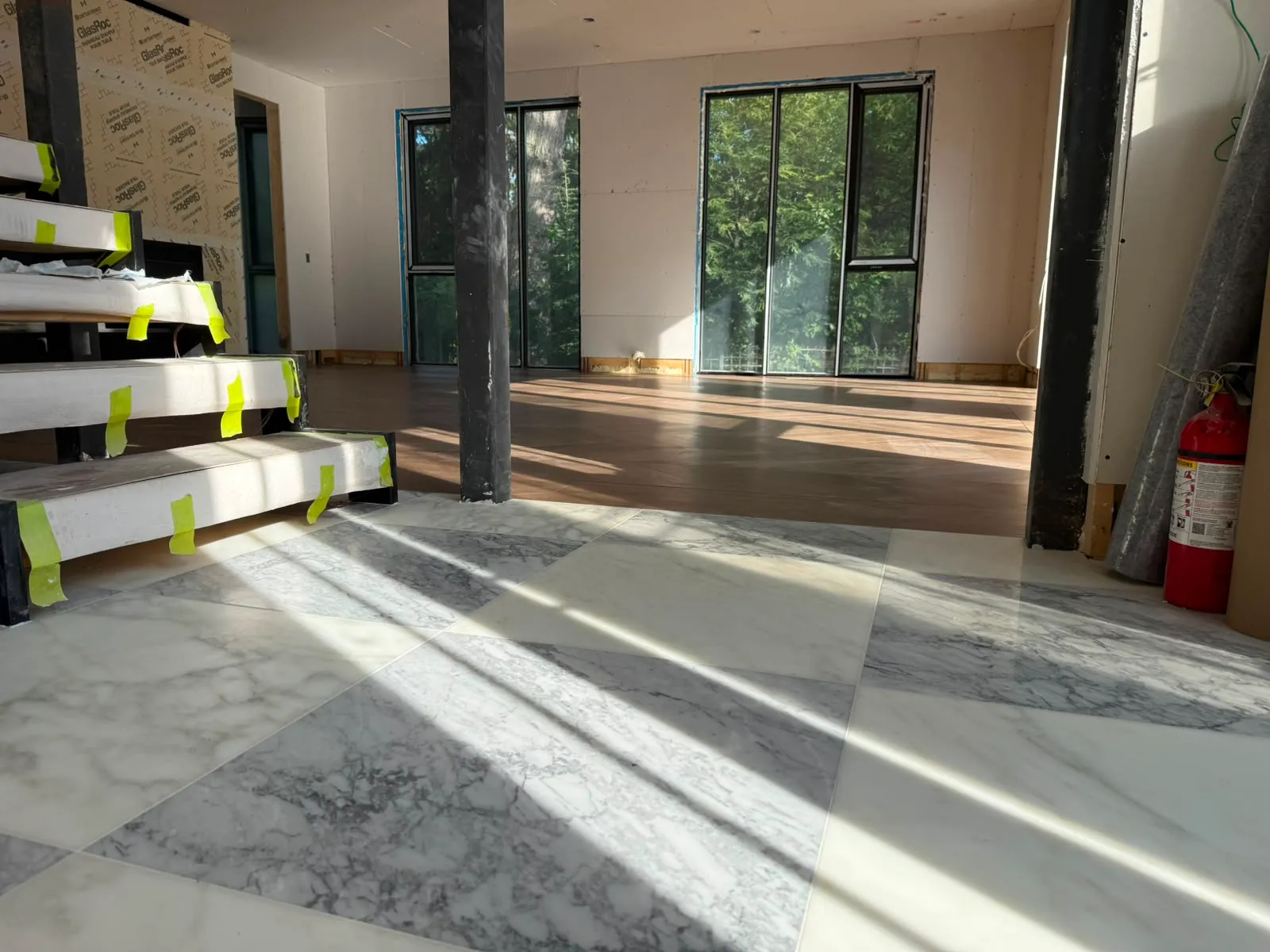 Stair treads wrapped in white protection and yellow tape beside a finished marble floor and tall windows