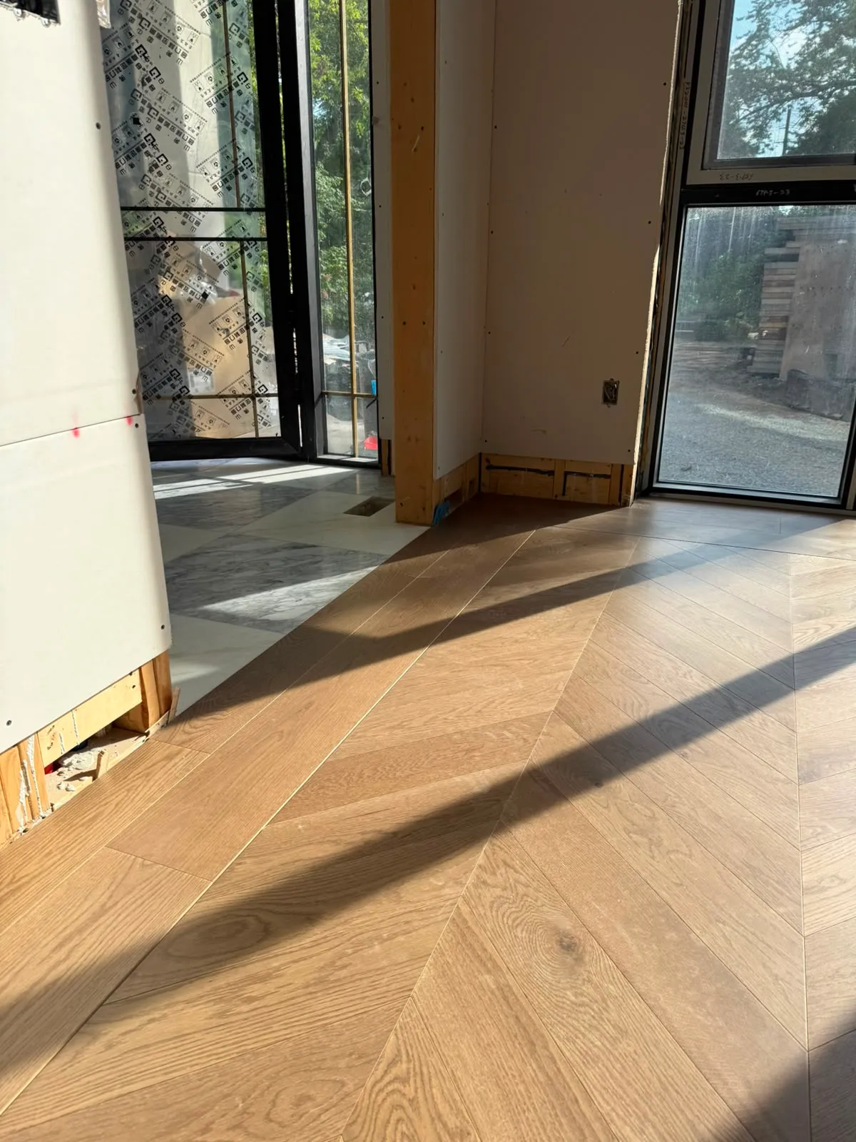 Chevron oak floor meeting grey and white marble at a doorway, with open framing at the base of the drywall