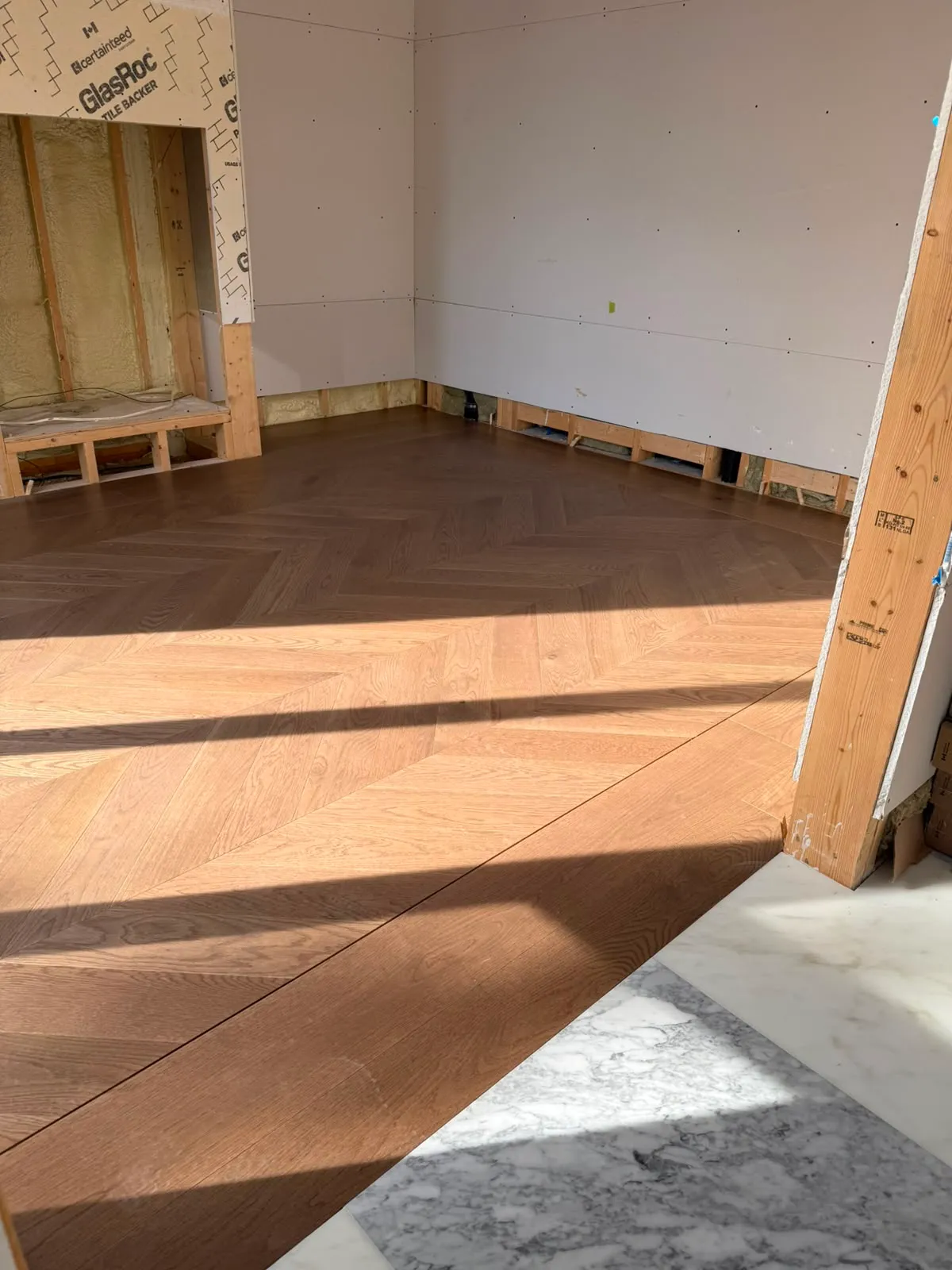 Chevron oak floor meeting marble at a doorway, a framed niche with spray foam insulation behind tile backer board, drywall not yet taped
