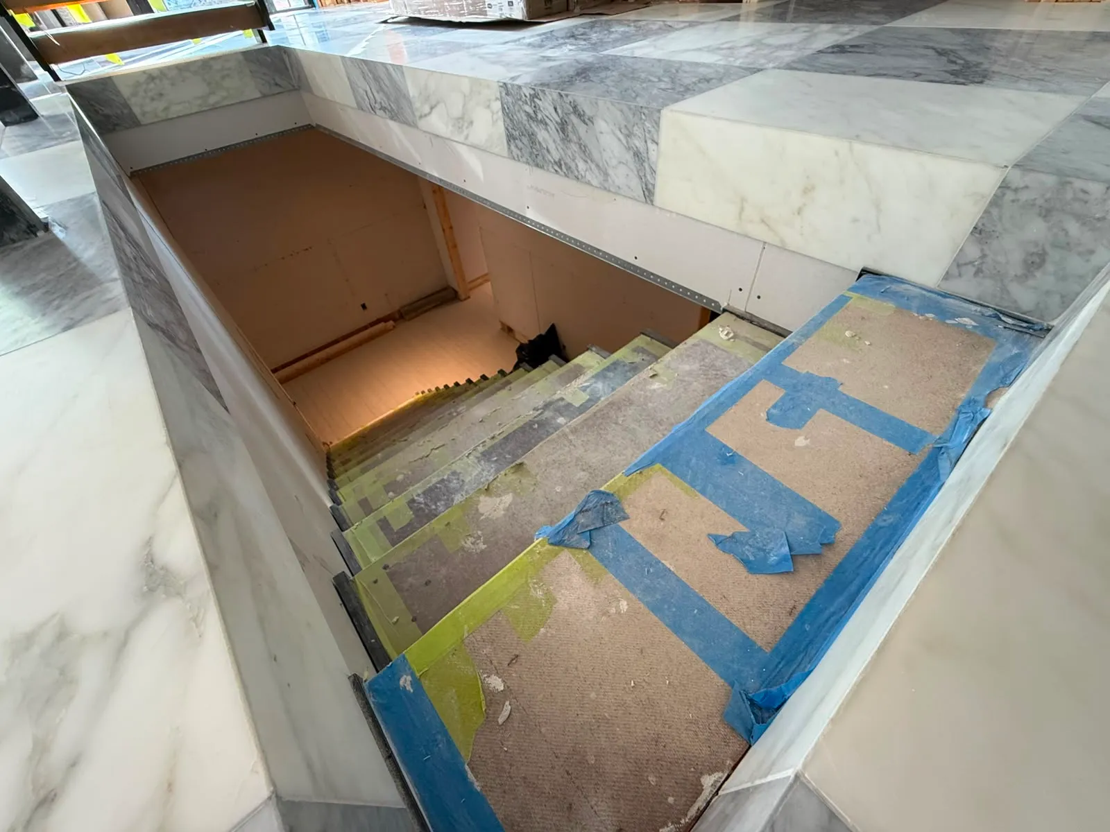 Looking down a staircase from a marble tiled floor: the treads are under protection board and tape, with drywalled walls on the floor below