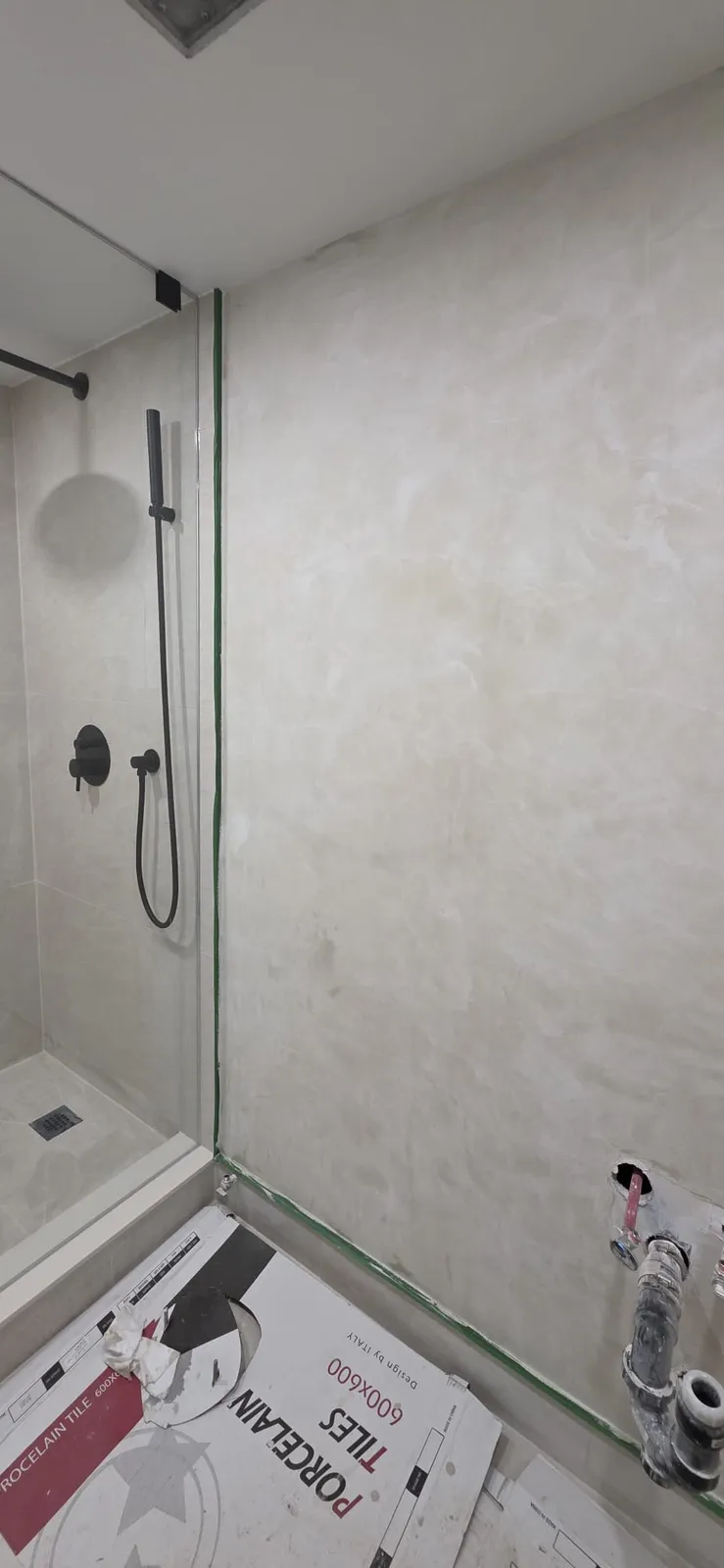Shower finished with large stone-look tile and a glass panel, beside it a tiled wall with water supply and drain pipes sticking out and a box of 600x600 porcelain tile on the floor