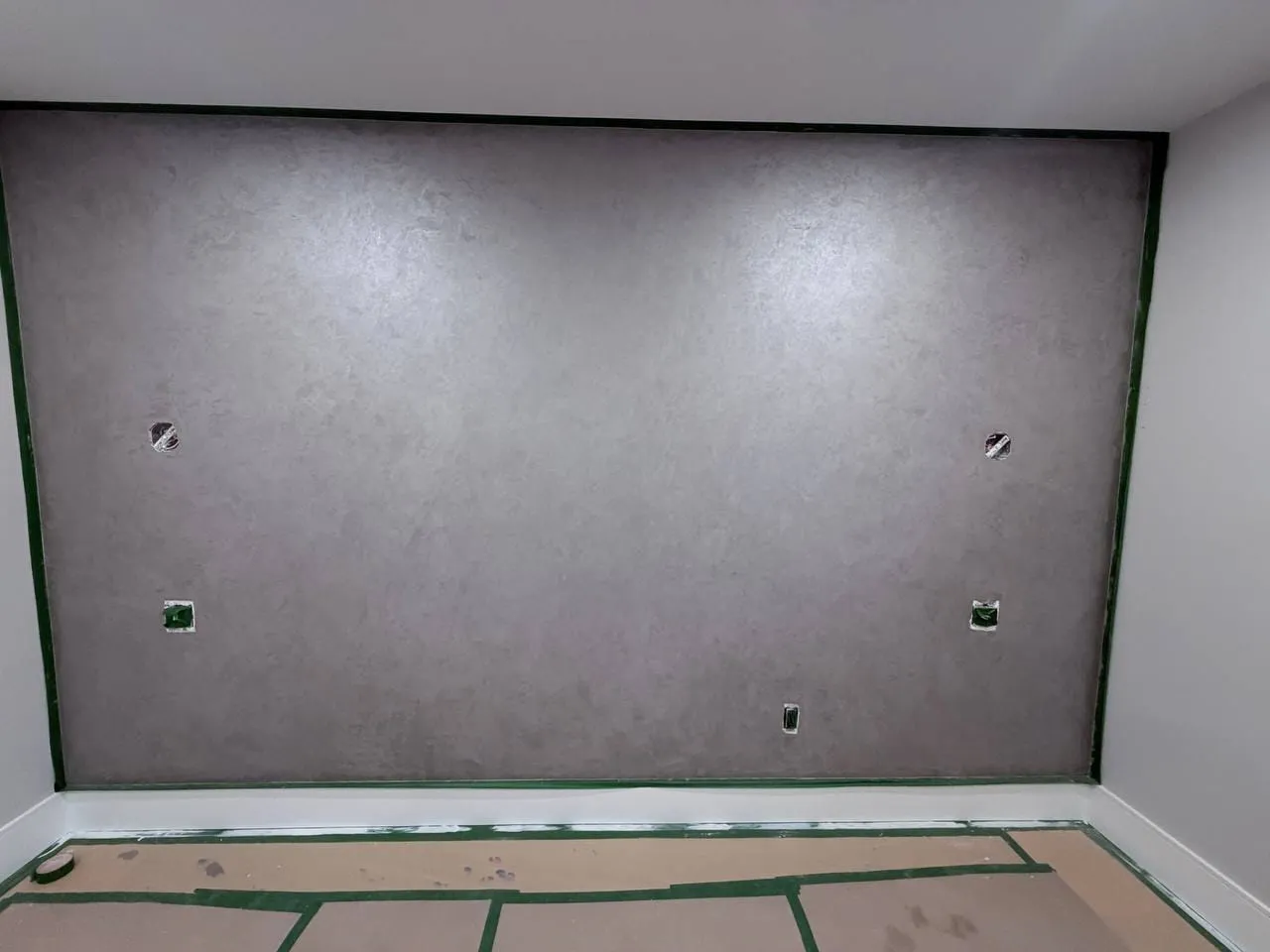 Grey plaster feature wall edged with green tape, with openings cut for wiring and a floor covered in protection paper