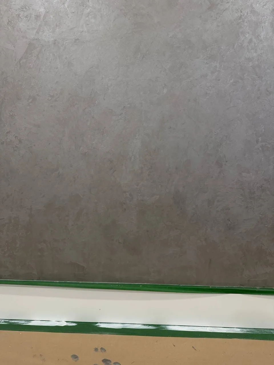 Grey troweled wall with green tape along the baseboard and paper covering the floor