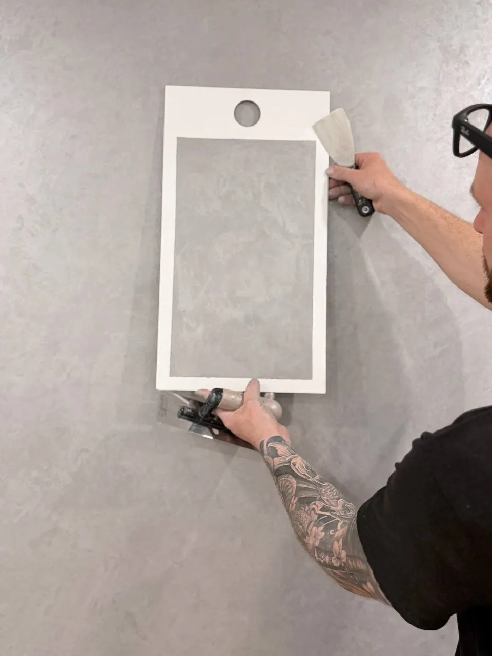 A hand holding a white sample board flat against a grey troweled wall