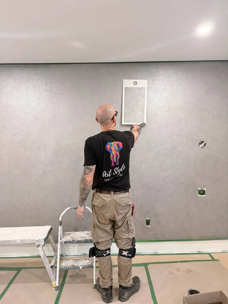A worker in an Art Style Construction shirt trowelling grey decorative plaster on a long wall, floor covered and taped