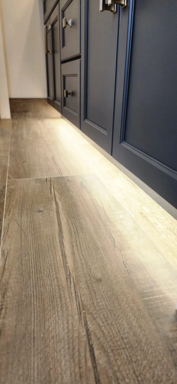 Close view of navy shaker vanity doors with brass pulls, a strip of light under the cabinet shining on wood-look plank tile