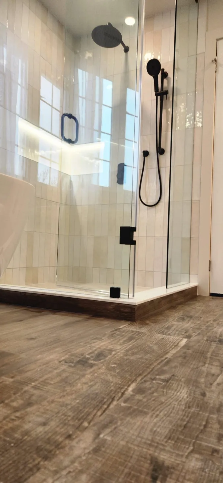Glass shower enclosure with black fixtures and a lit niche in the white tile, wood-look floor tile in front