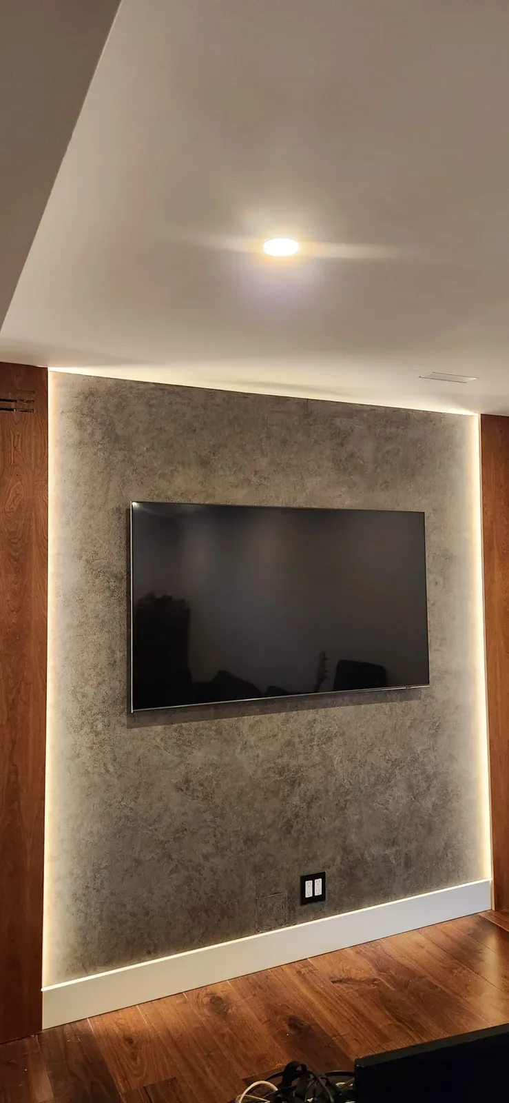 Flat-screen TV mounted on a grey plaster wall with LED light along its edges, a double outlet below and a wood floor