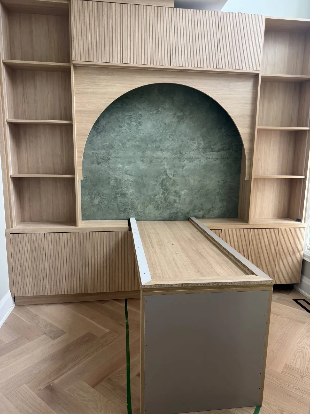 Oak built-in shelving and fluted cabinets around an arched niche finished in green textured plaster, with a desk being installed
