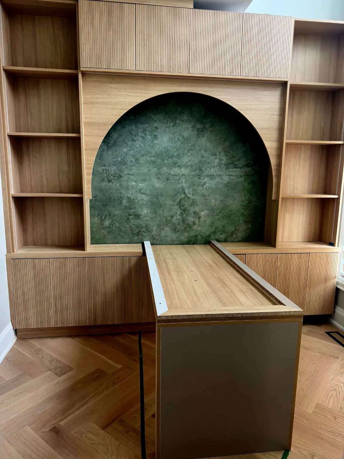 Oak built-in with open shelves and fluted panels around an arched opening lined with a green stone-look panel, over a herringbone floor
