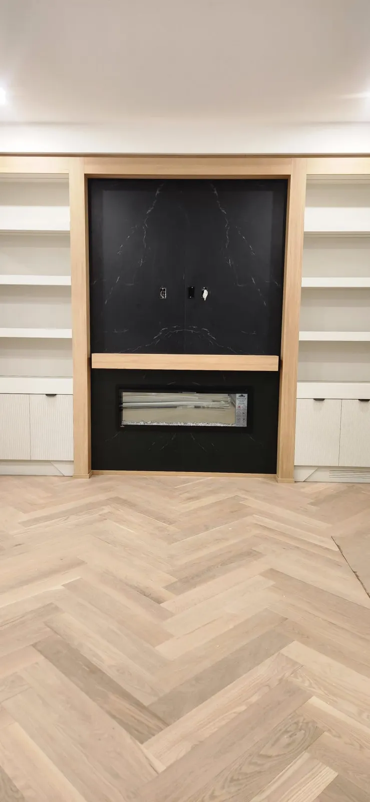 Black stone fireplace wall framed in light oak, with white shelving on both sides and a light herringbone floor