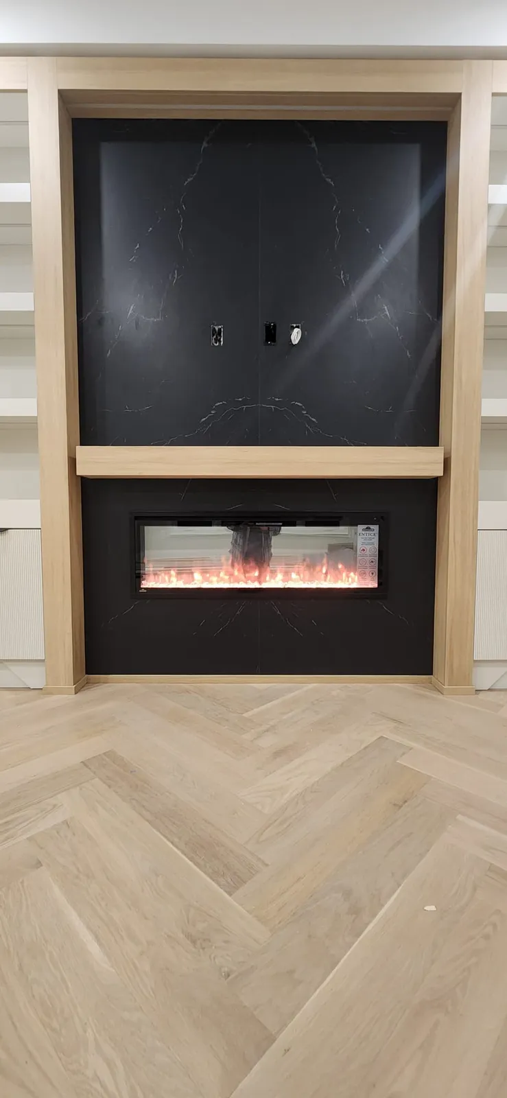 Media wall with a black stone-look panel above a linear electric fireplace, framed in light oak, over a light herringbone floor