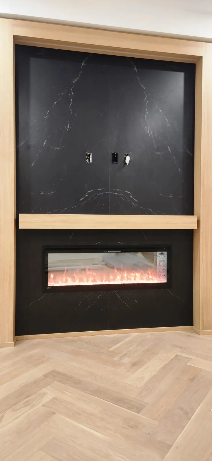 Media wall with a black marble-look slab, an electric fireplace insert and a light oak frame and mantel above a herringbone oak floor