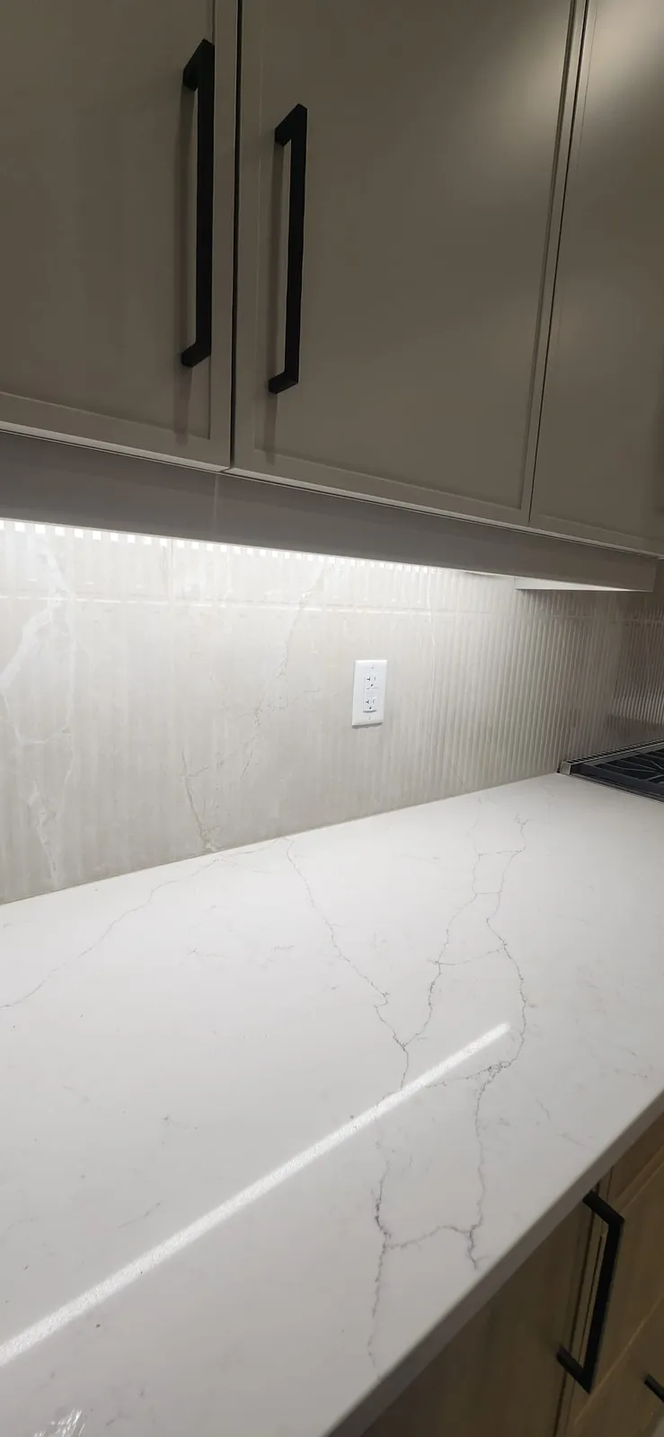 White veined counter meeting a fluted tile backsplash with an outlet, under grey cabinets with black handles