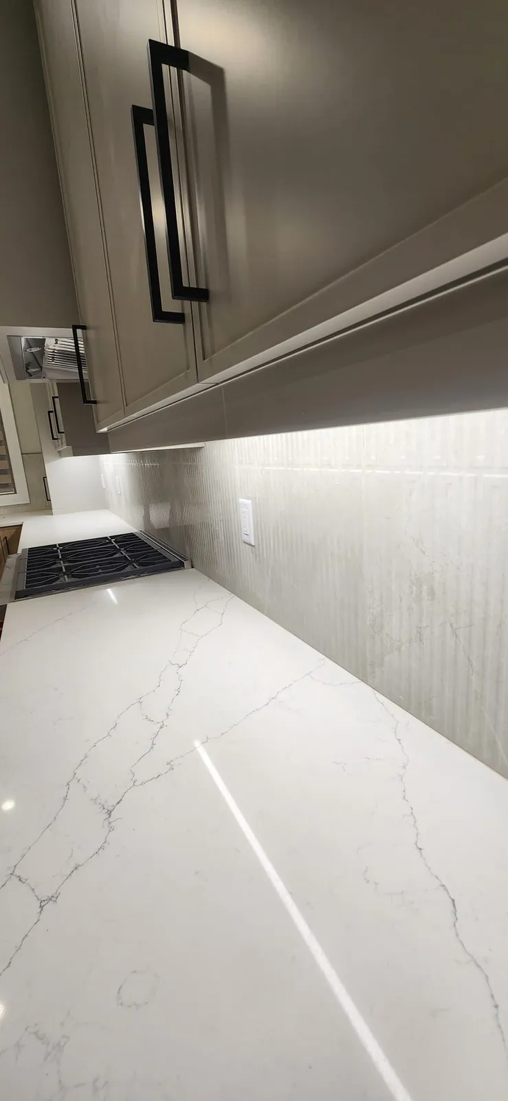 View along a fluted tile backsplash with a white outlet and under-cabinet light, above a white veined counter leading to a gas range