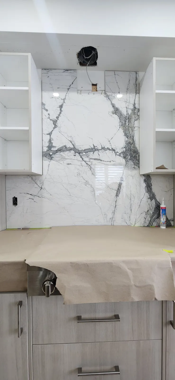 White marble-look slab backsplash being fitted between open upper cabinets, with the counter covered in paper and the hood duct opening still exposed