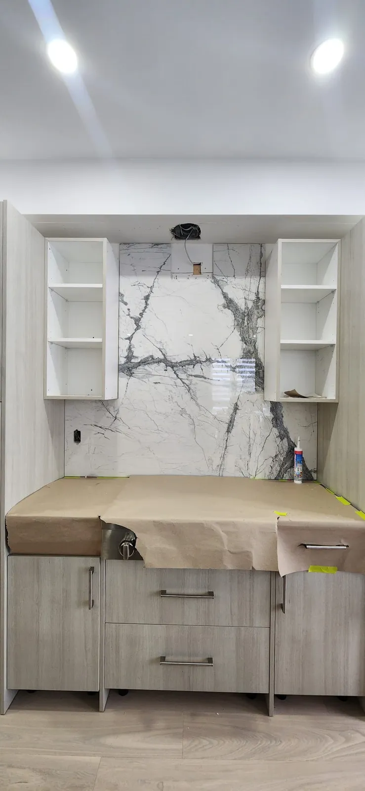 Kitchen wall mid-installation: a white slab backsplash with grey veining between two open shelf boxes, an outlet cut-out at left, an open wiring box above, the counter under brown paper and a caulking tube
