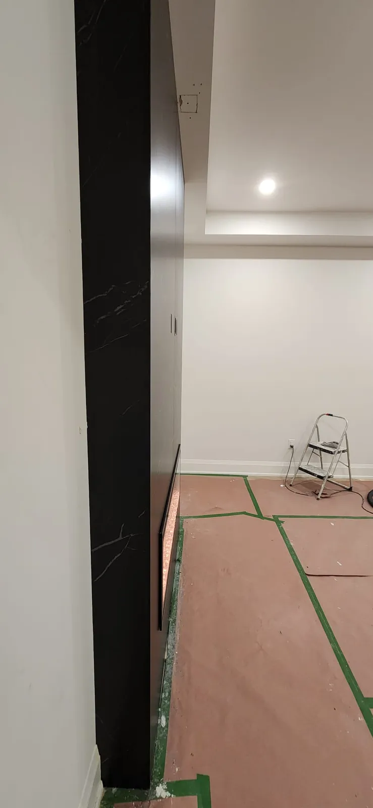 A stepped white ceiling with a small square cut-out still open near its edge and a pot light, plain white walls, a black veined panel wall on the left, the floor under brown paper with green tape and a step stool by the wall