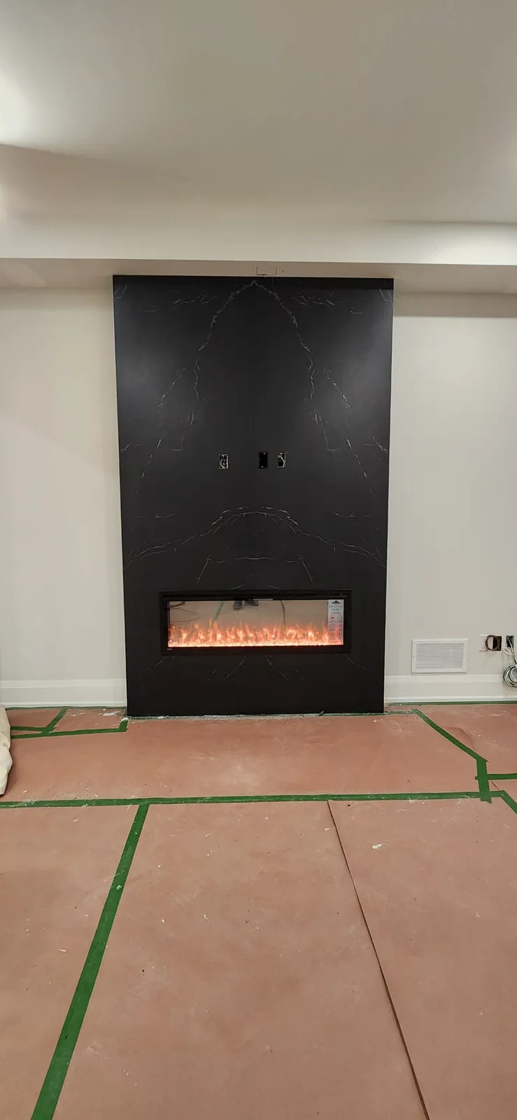 Black veined slab wall with a lit electric fireplace, the floor still covered with paper and green tape