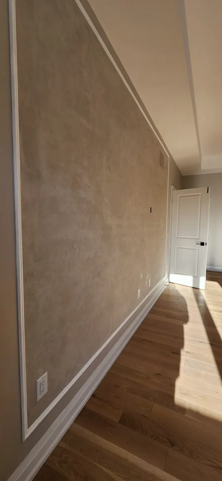 Long wall with beige troweled panels framed by white moulding, painted beige walls, white baseboard and a white panel door