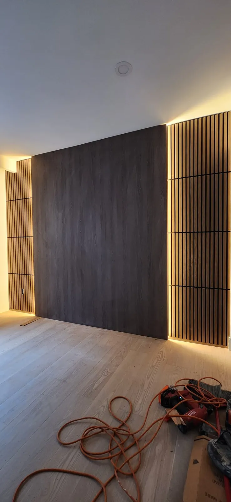 Dark wood-look panel between two slatted wall sections with warm LED light behind, extension cords on the new floor