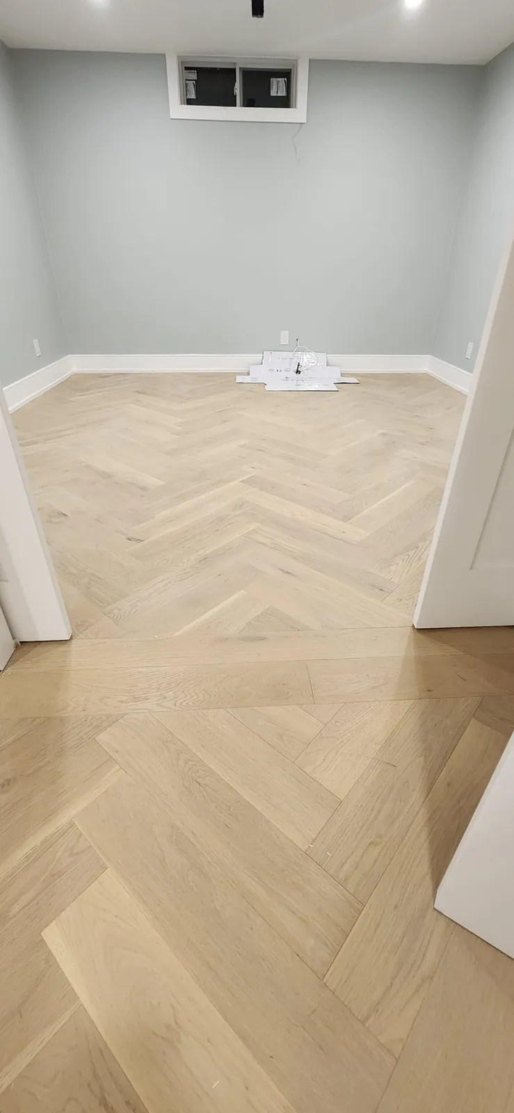 Finished basement bedroom with pale grey walls, white baseboards, a small window high on the wall and a light herringbone oak floor